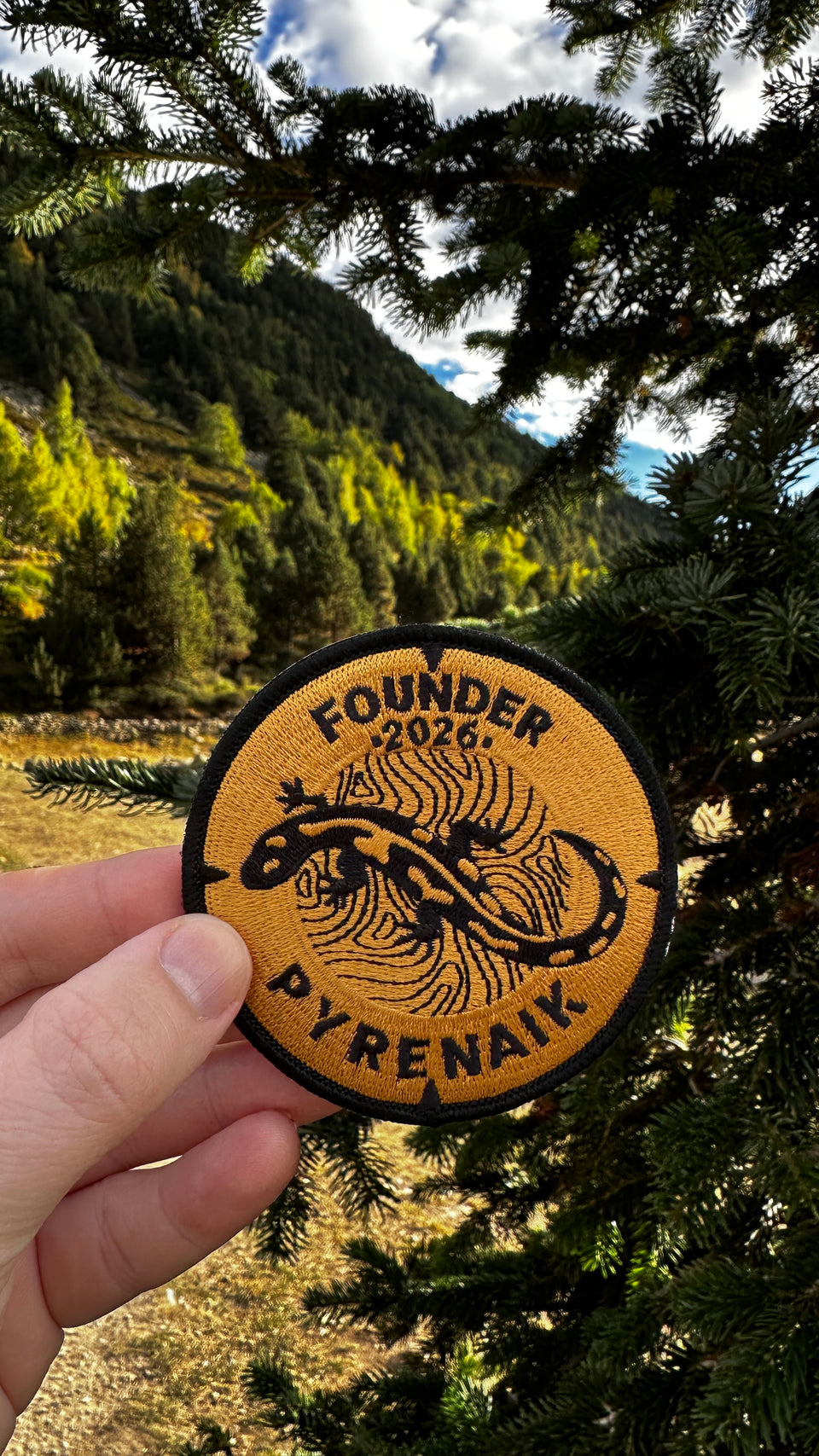 Parche Pyrenaik Founders Edition 2026 bordado en negro y dorado, sostenido en la montaña